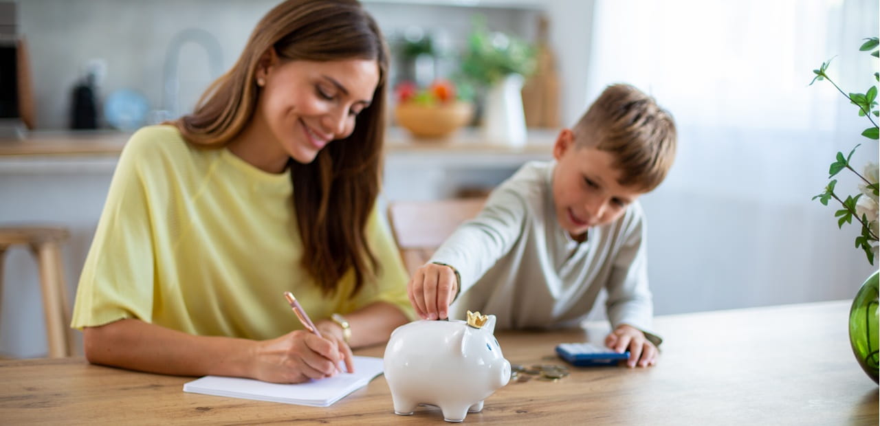Mother and son sitting at a table, writing and placing coins into a white piggy bank.