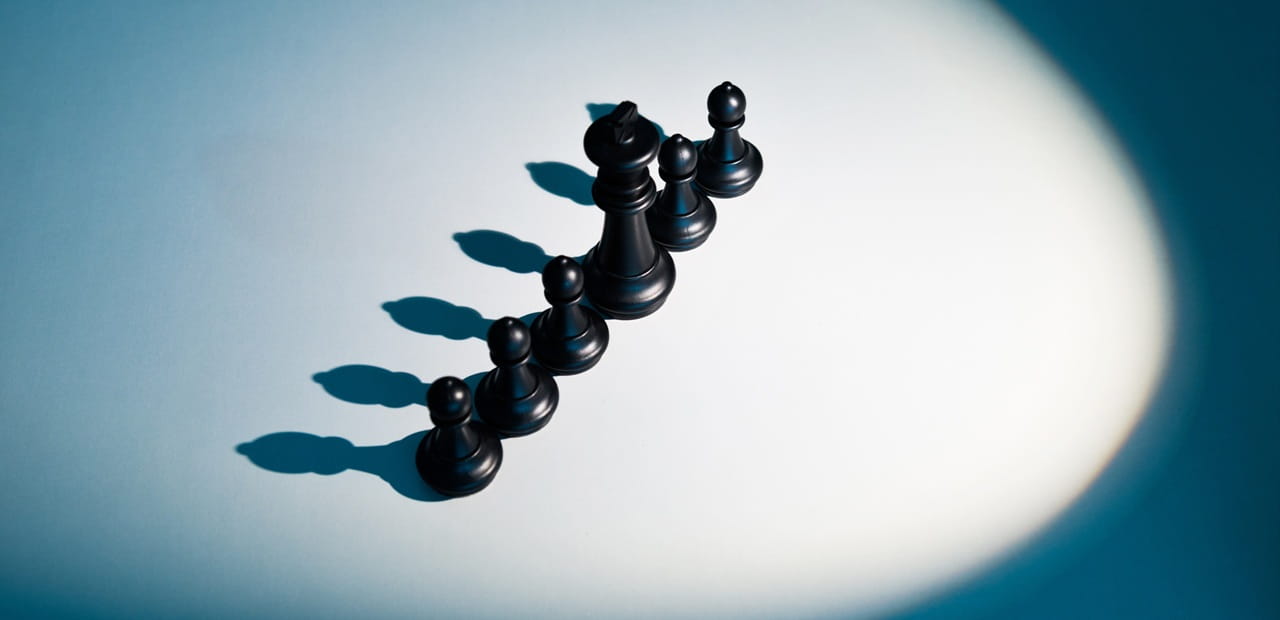 Black chess pieces arranged in a diagonal line on a light surface, casting long shadows