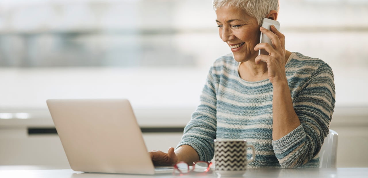 Person working at a laptop while talking on a mobile phone, with a coffee mug on the table.