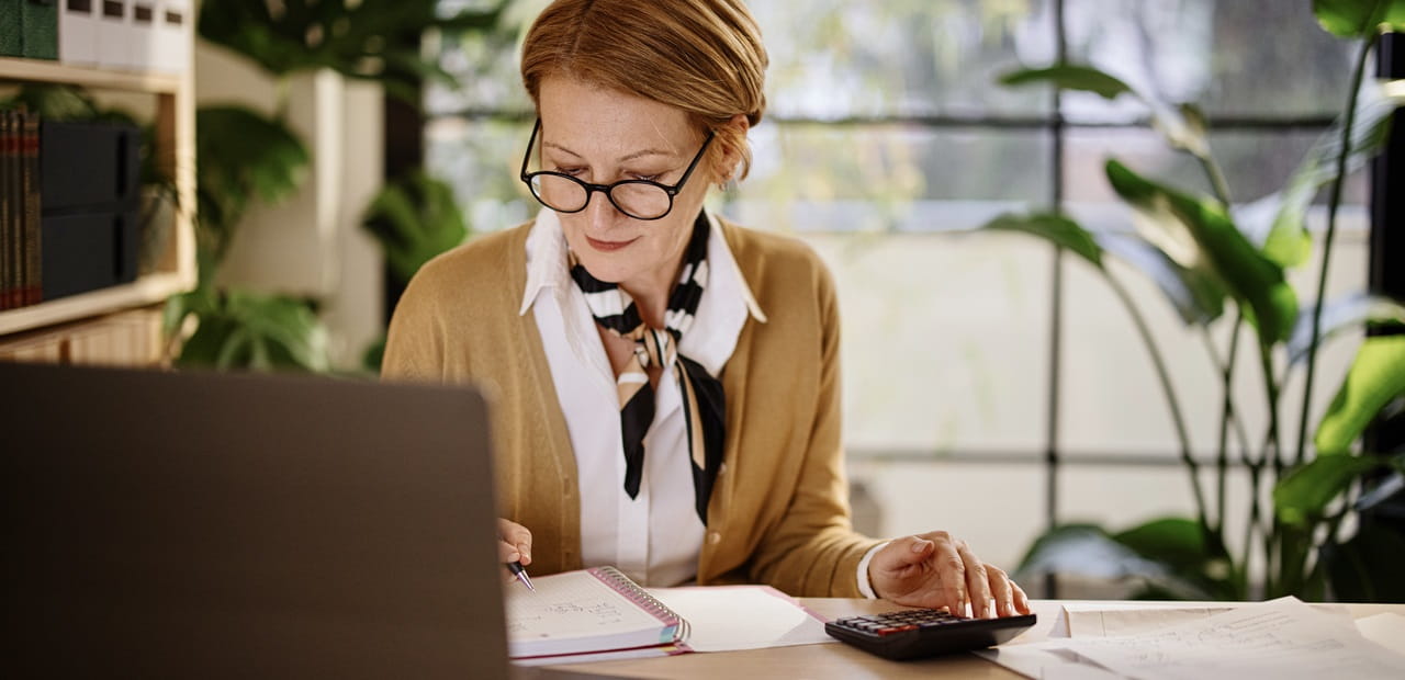 A person sits at a desk in a bright office space with large windows and green plants, working on a laptop while writing in a notebook and using a calculator surrounded by papers.