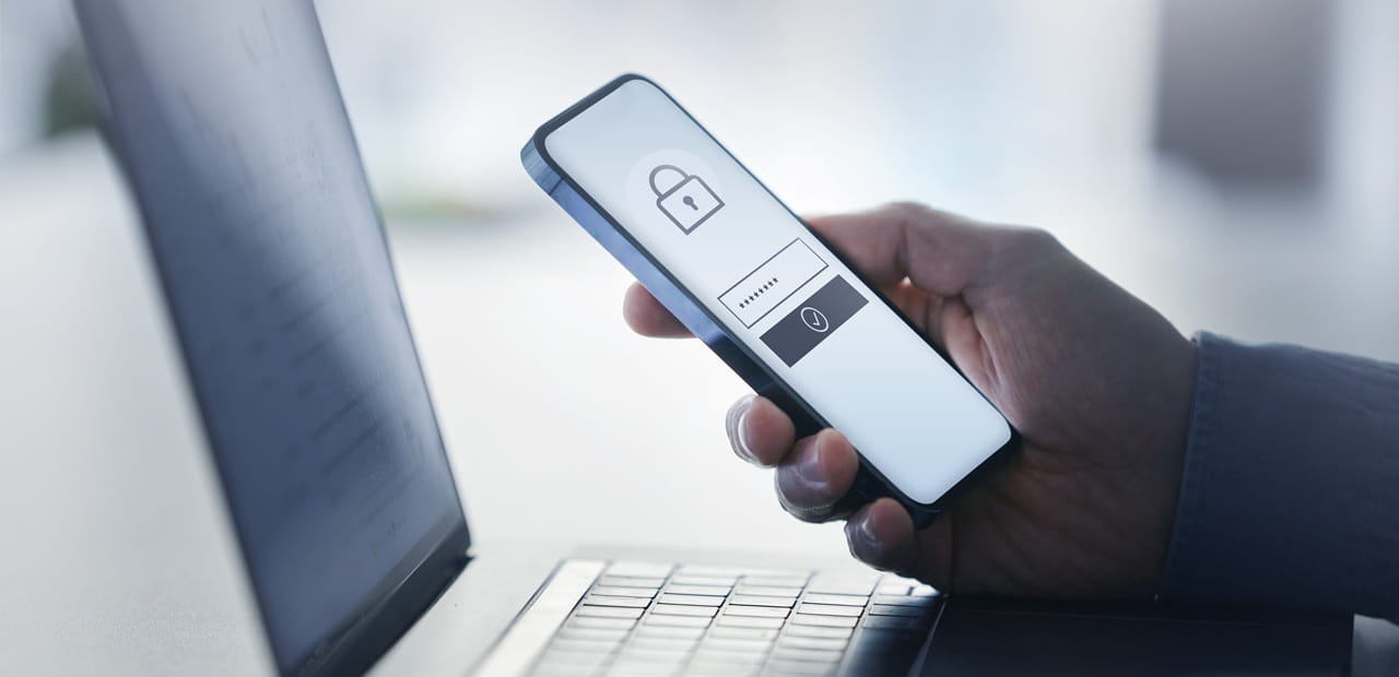 Hand holding a smartphone with a lock icon beside a laptop, illustrating cybersecurity and secure authentication.