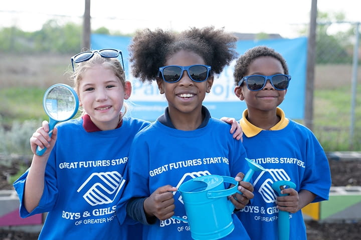 Three diverse elementary age Boys and Girls Clubs of America members smiling at camera holding gardening tools